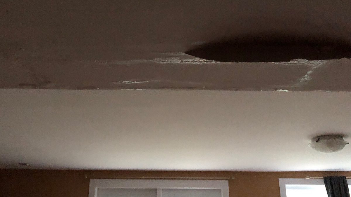 Dark water stain on a home ceiling above a window, a sign of a ceiling leak