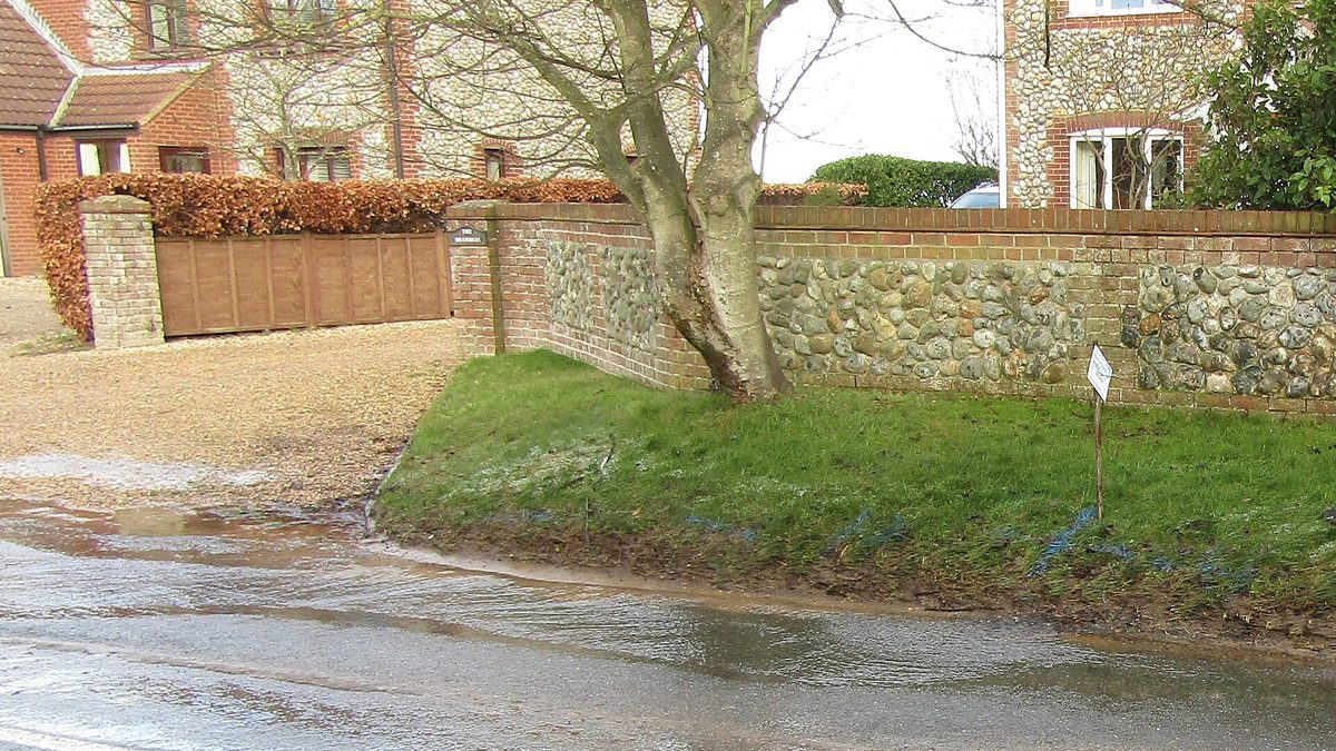 Burst water main spilling water across Mundesley Road in Trimingham, Norfolk, beside brick houses