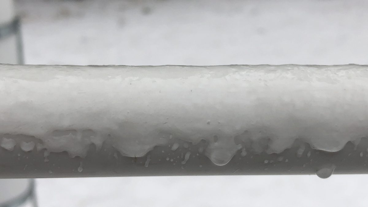 Close-up of a frozen PVC water pipe heavily coated with thick glaze ice, with icicles and ice accumulation