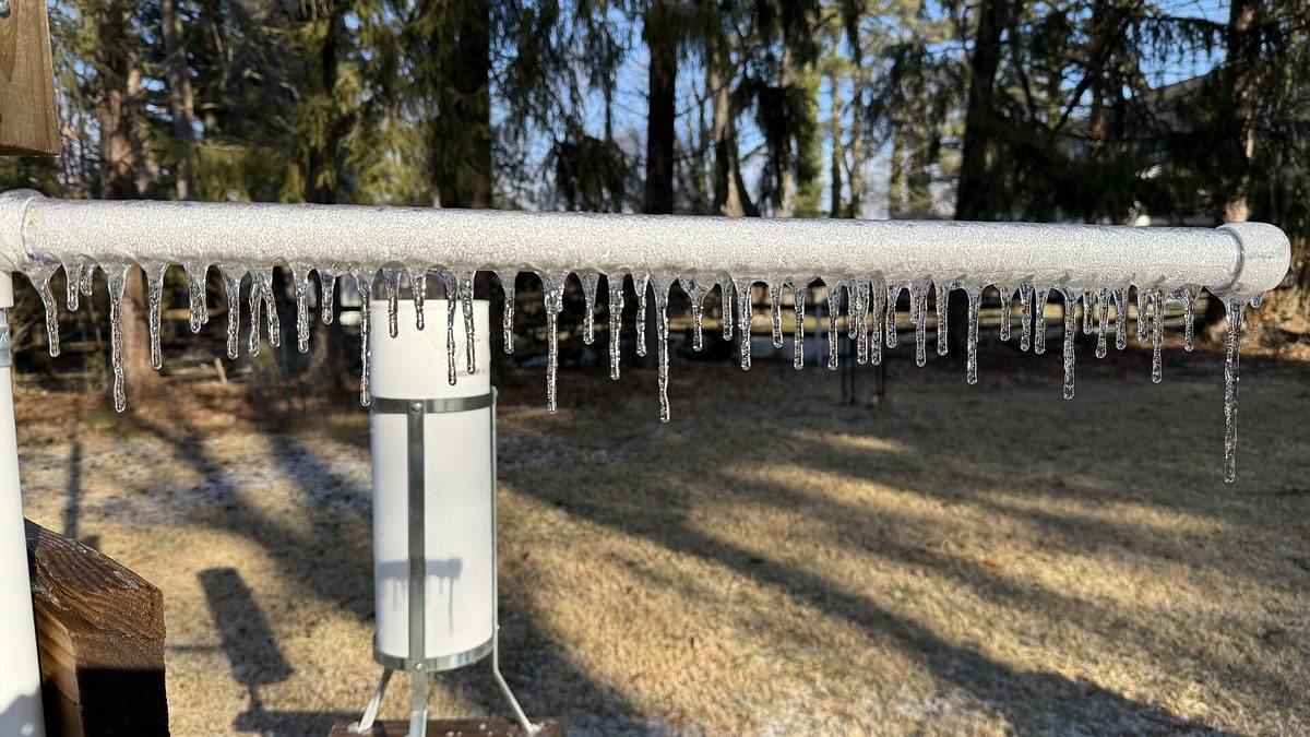 A frozen outdoor water pipe in a backyard heavily coated in thick glaze ice and hanging icicles after freezing rain