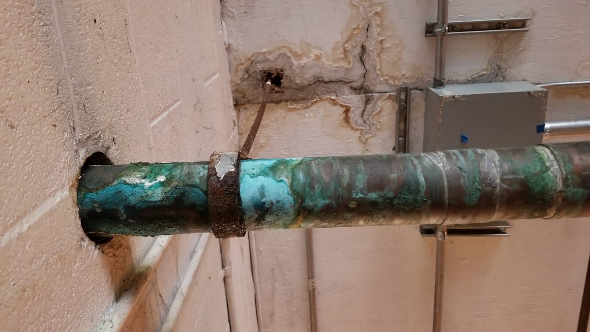 Pinhole leak corrosion on a copper water pipe, with green and white mineral staining along a basement wall