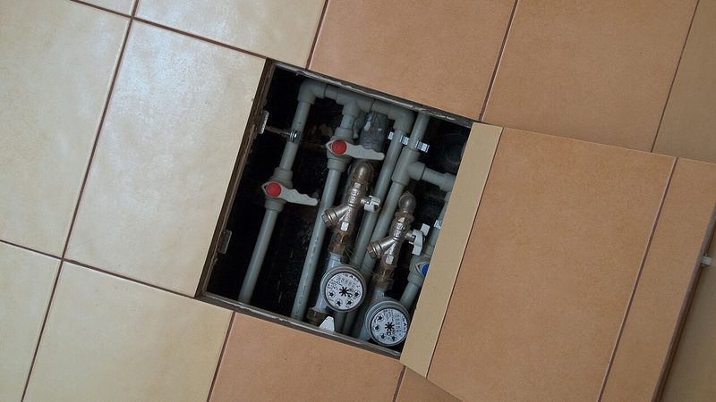 Open bathroom floor access panel showing shutoff valves, water supply pipes, and water meters, the kind of plumbing behind the wall or floor when a pipe bursts in a bathroom