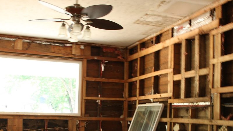 Interior wall opened down to the studs after a burst pipe caused water damage, showing exposed framing, a window, and a ceiling fan