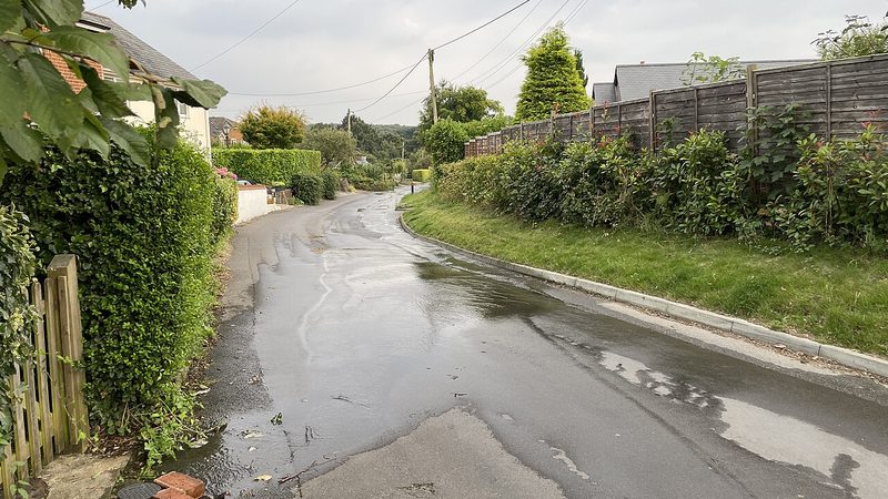 Water from a leaking water main flooding a residential street beside houses and hedges